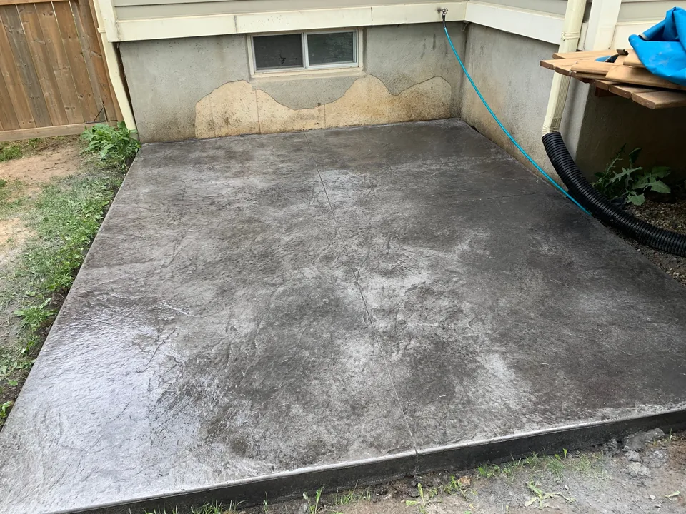A rectangular dark textured concrete patio sits between a house foundation and a timber fence.
