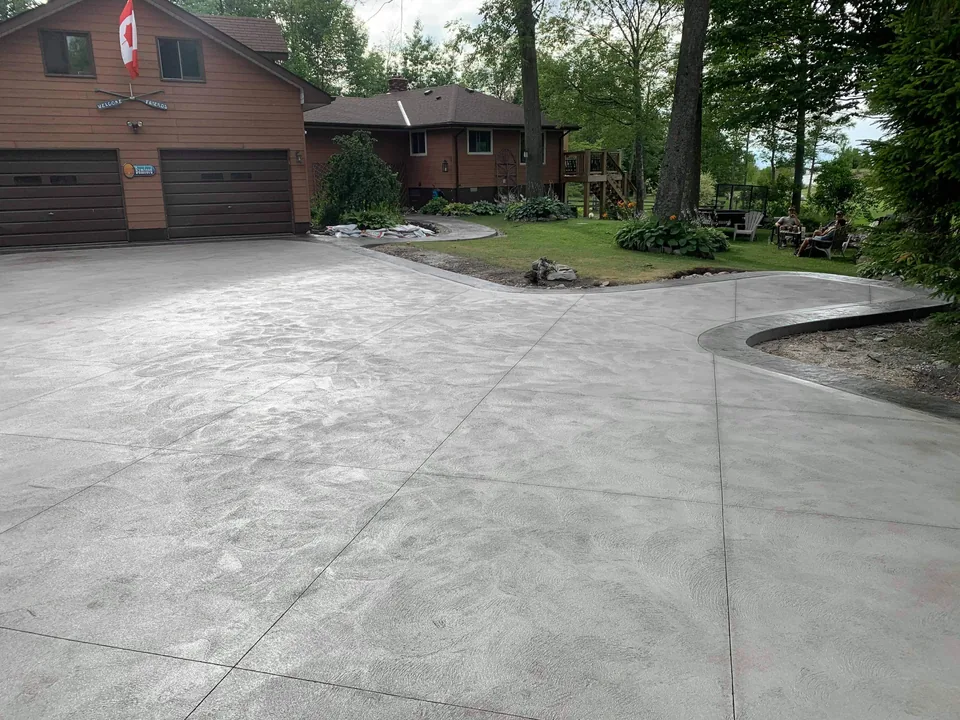 A broad concrete driveway meets a curved path beside a lawn and mature trees.