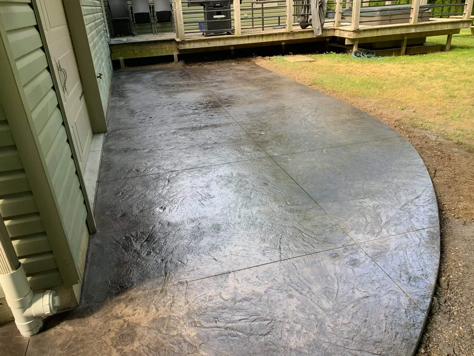 A dark textured concrete patio with a rounded outer edge sits beside a house and raised timber deck.