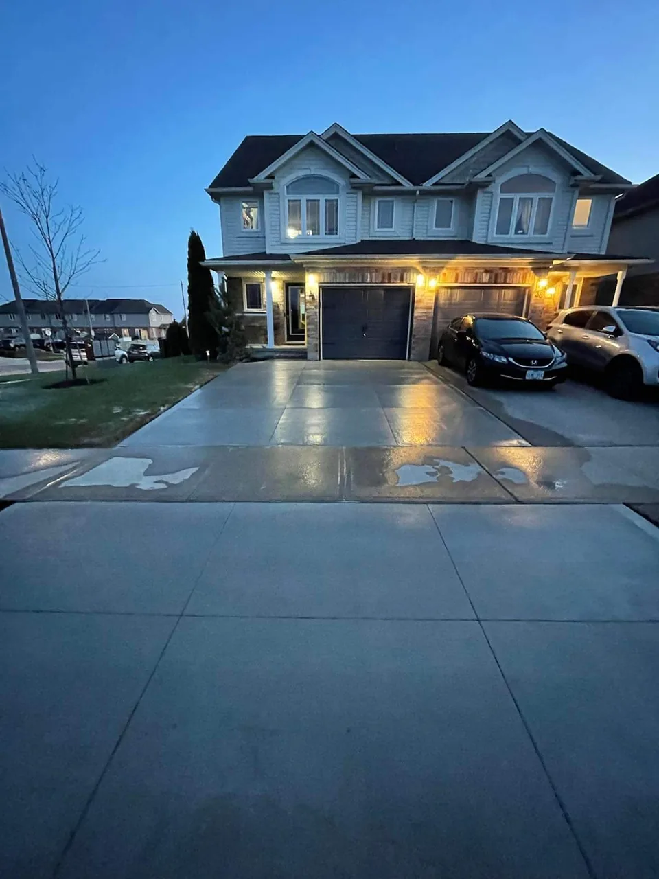 A concrete driveway reflects porch lights in front of a two-storey home at dusk.