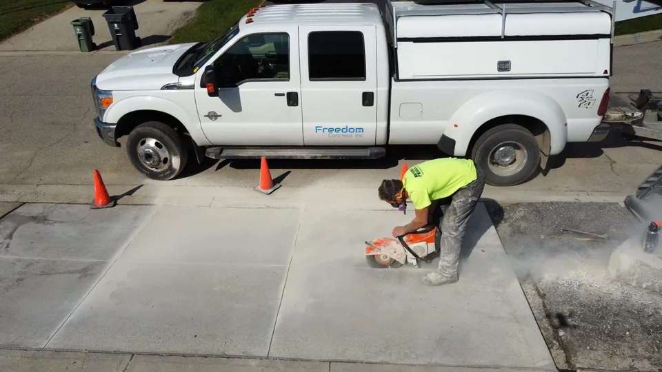 A crew member uses a concrete saw on a driveway beside a parked work truck and traffic cones.