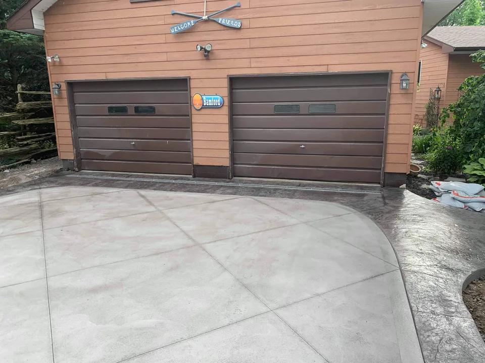 A concrete apron in front of two garage doors has diagonal joints, a darker edge, and a drainage channel.