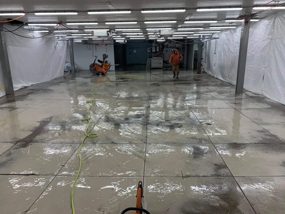 A crew member and floor equipment stand on a wet concrete floor marked with straight joints.