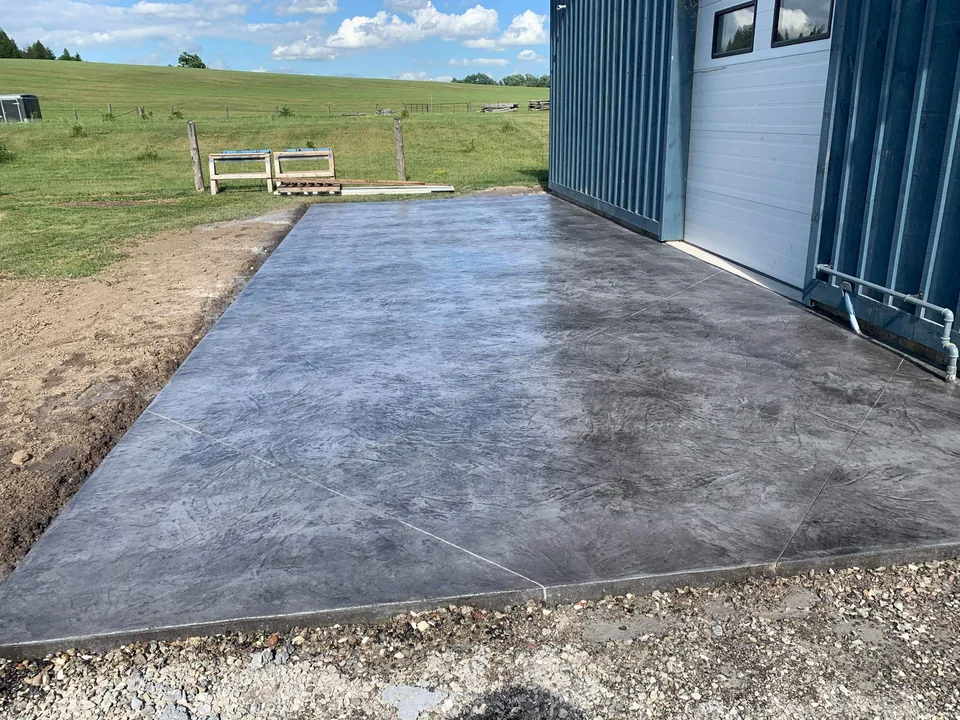 A rectangular dark textured concrete apron extends from a metal-sided building toward a grassy field.