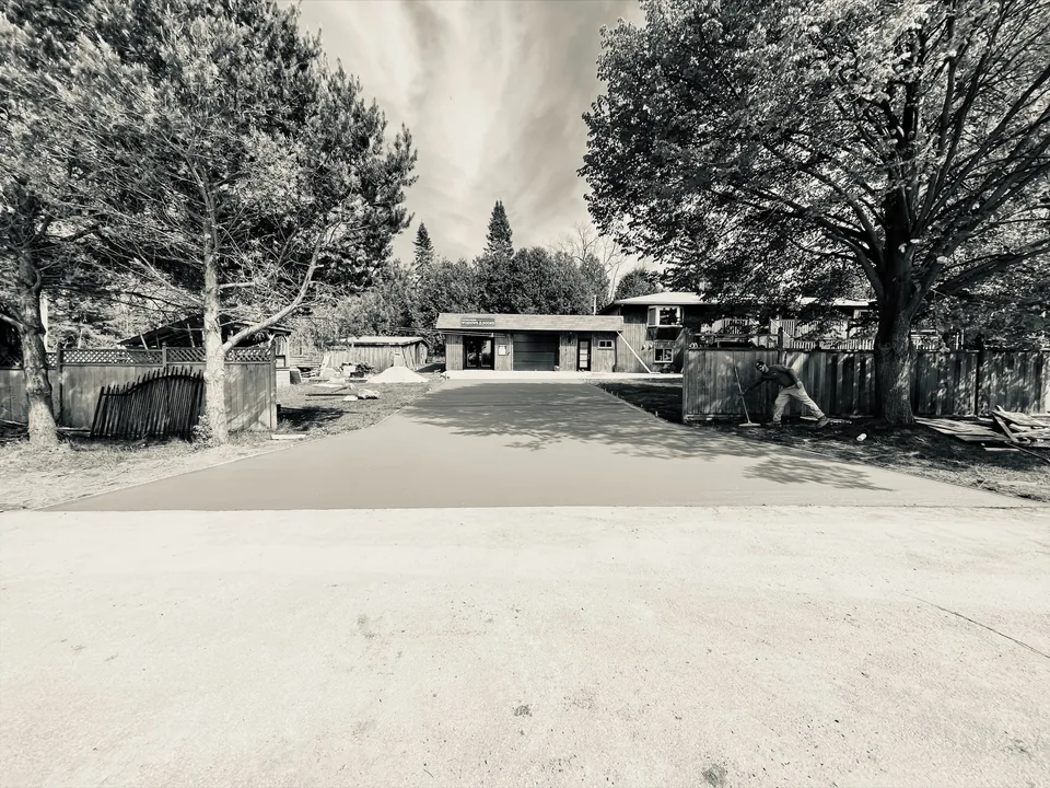 A monochrome photograph of a wide concrete driveway leading between trees to a garage.